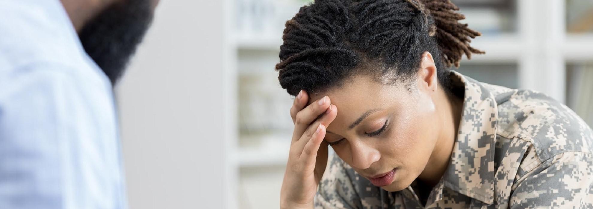 Woman in military uniform holding her head in pain as doctor in white coat looks at her.