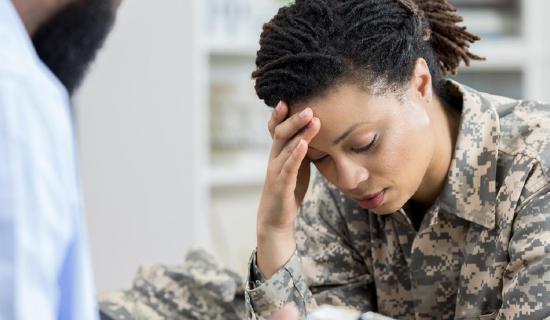 Woman in military uniform holding her head in pain as doctor in white coat looks at her.